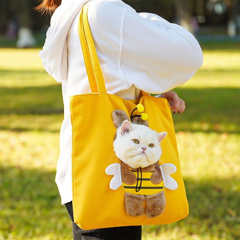A person outdoors carrying a bright yellow tote bag with a plush cat dressed as a bee on the front. The person is wearing a white hoodie, and the background shows a blurred grassy park setting.