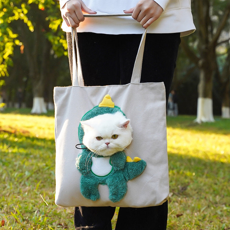 Person holding a tote bag with a cat in a dinosaur costume plushie on a grassy outdoor background